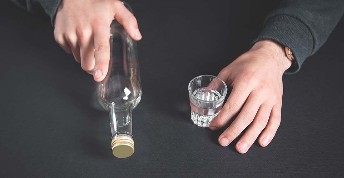 Руки держат бутылку алкоголя и полную рюмку с бесцветной жидкостью